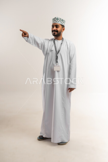 اشارات تحدد هذا المكان (هنا)، الاشارة إلى شيء ما، تعابير التحديد والتأكيد، بورتريه لرجل عربي خليجي عماني مبتسم يرتدي الدشداشه والكمة يشير بإصبع السبابة وينظر الى شيء ما بإيماءات السعادة والسرور، الجسم الكامل بالطول، خلفية بيضاء