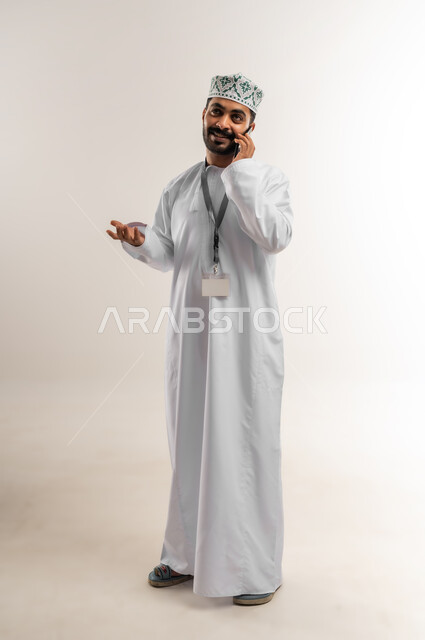 ارتداء البطاقة التعريفي، دمج التكنولوجيا والاجهزة الحديثة في الحياة اليومية، بورتريه لرجل عربي خليجي عماني يرتدي الدشداشه والكمة يقوم بإجراء مكالمة هاتفية باستخدام الهاتف المحمول، التواصل مع الاهل والاصدقاء، دردشة ومحادثات وتعارف، الجسم بالطول الكامل، خلفية بيضاء