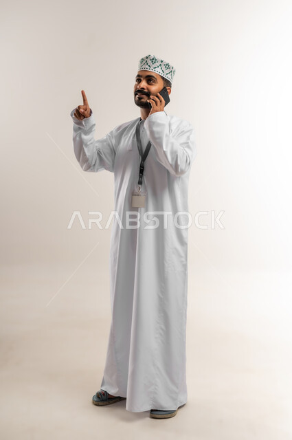 ارتداء البطاقة التعريفي، دمج التكنولوجيا والاجهزة الحديثة في الحياة اليومية، بورتريه لرجل عربي خليجي عماني يرتدي الدشداشه والكمة يقوم بإجراء مكالمة هاتفية باستخدام الهاتف المحمول، التواصل مع الاهل والاصدقاء، دردشة ومحادثات وتعارف، الجسم بالطول الكامل، خلفية بيضاء