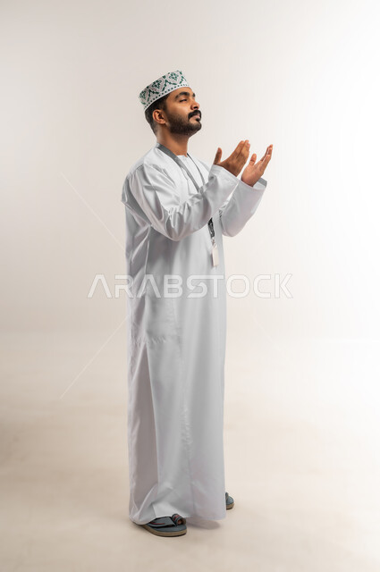 الإكثار من الدعاء في شهر رمضان المبارك، الإجتهاد في العبادة والحفاظ على الطاعات، بورتريه لشاب عربي خليجي عماني يرتدي الدشداشة والكمة يرفع يديه ويدعو الله بإيماءات التوسل والتضرع، صورة الجسم بالطول الكامل, خلفية بيضاء