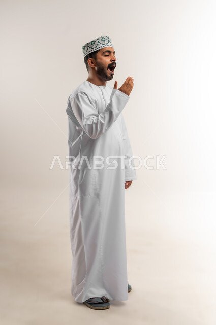 الشعور بالتعب والحاجة الى النوم والراحة, الإحساس بالكسل والخمول و الإرهاق، بورتريه لشاب عربي خليجي عماني يرتدي الدشداشة و الكمة يضع يده على فمه بإيماءات التثاؤب والنعاس، صورة الجسم بالطول الكامل, خلفية بيضاء