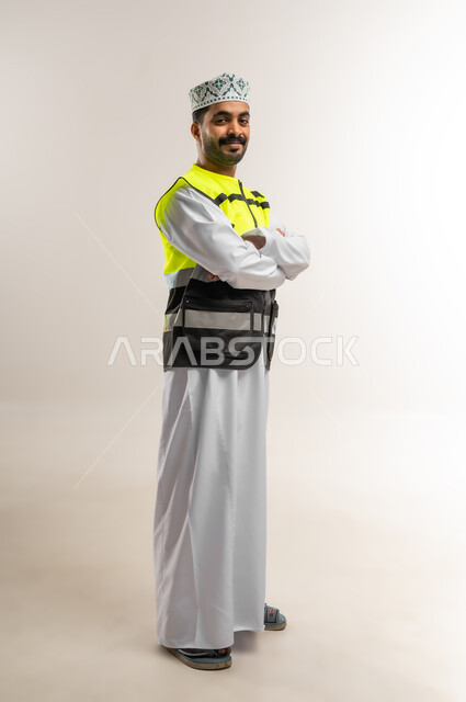 Concept of engineering and architectural construction, portrait of an Arab Gulf Omani engineer wearing a dishdasha, a keffiyeh, and a protective vest, standing with crossed hands looking at the camera with an expression of pleasure and happiness, profession and job for young people in the Sultanate of Oman, working in the engineering sector, full body, white background