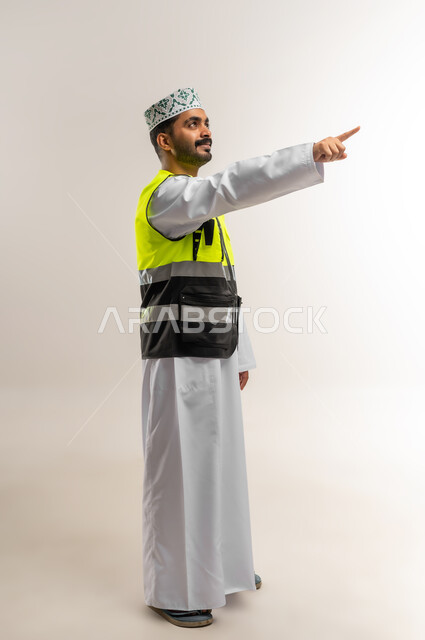 الاشارة الى شي ما، مفهوم الهندسة والانشاءات المعمارية، بورتريه لمهندس عربي خليجي عماني يرتدي الدشداشة والكمة وسترة الحماية يشير لمكان ما، مهنة ووظيفة للشباب في سلطنة عمان، العمل في القطاع الهندسي، الجسم بالطول الكامل، خلفية بيضاء