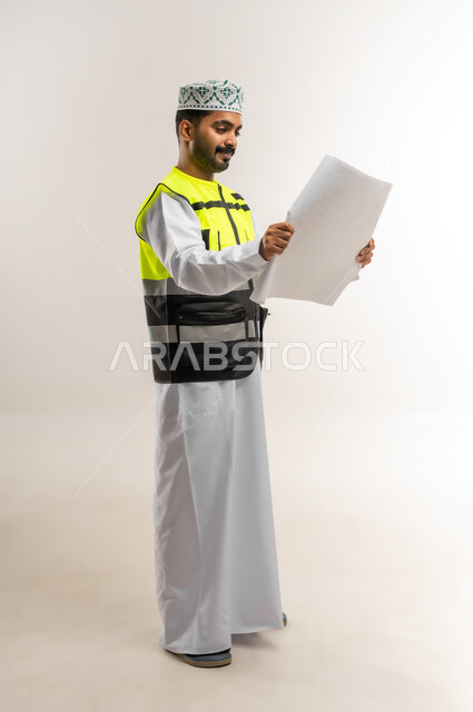 Working in the engineering sector, the concept of engineering and architectural construction, a portrait of an Arab Gulf Omani engineer wearing a dishdasha, a keffiyeh, and a protective vest, holding a construction plan in his hand, a profession and job for young people in the Sultanate of Oman, full-length body, white background