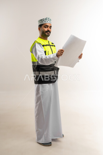 العمل في القطاع الهندسي، مفهوم الهندسة والانشاءات المعمارية، بورتريه لمهندس عربي خليجي عماني يرتدي الدشداشة والكمة وسترة الحماية يمسك بيده مخطط البناء، مهنة ووظيفة للشباب في سلطنة عمان، الجسم بالطول الكامل، خلفية بيضاء