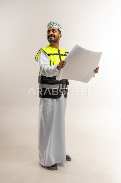 Working in the engineering sector, the concept of engineering and architectural construction, a portrait of an Arab Gulf Omani engineer wearing a dishdasha, a keffiyeh, and a protective vest, holding a construction plan in his hand, a profession and job for young people in the Sultanate of Oman, full-length body, white background
