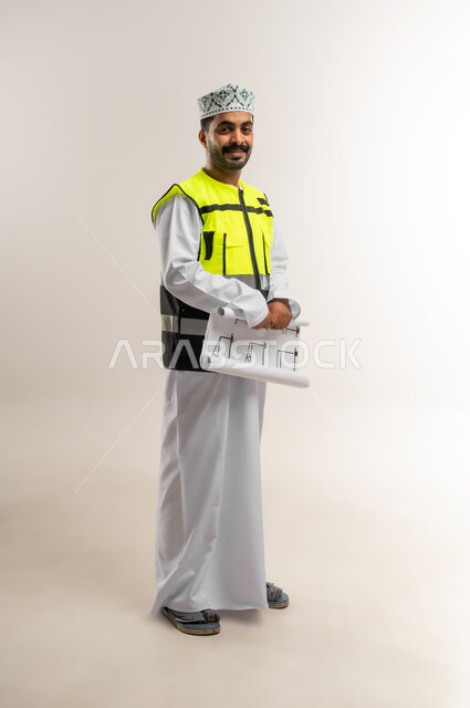 العمل في القطاع الهندسي، مفهوم الهندسة والانشاءات المعمارية، بورتريه لمهندس عربي خليجي عماني يرتدي الدشداشة والكمة وسترة الحماية يمسك بيده مخطط البناء، مهنة ووظيفة للشباب في سلطنة عمان، الجسم بالطول الكامل، خلفية بيضاء