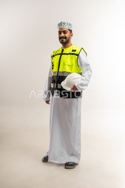 Working in the engineering sector, the concept of engineering and architectural construction, a portrait of an Arab Gulf Omani engineer wearing a dishdasha, a keffiyeh, and a protective vest, holding a white helmet with an expression of pleasure, a profession and job for young people in the Sultanate of Oman, full body, white background