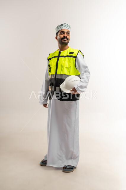 Working in the engineering sector, the concept of engineering and architectural construction, a portrait of an Arab Gulf Omani engineer wearing a dishdasha, a keffiyeh, and a protective vest, holding a white helmet with an expression of pleasure, a profession and job for young people in the Sultanate of Oman, full body, white background