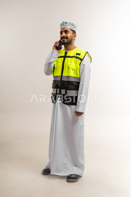 الرد على الاتصال الهاتفي, العمل في القطاع الهندسي، مفهوم الهندسة والانشاءات المعمارية، بورتريه لمهندس عربي خليجي عماني يرتدي الدشداشة والكمة وسترة الحماية يجري مكالمة عبر الهاتف المحمول، مهنة ووظيفة للشباب في سلطنة عمان، الجسم بالطول الكامل، خلفية بيضاء