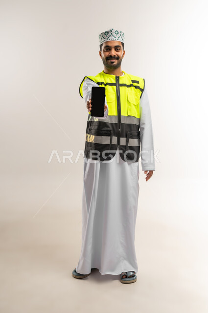 Empty black screen, working in the engineering sector, concept of engineering and architectural construction, portrait of an Arab Gulf Omani engineer wearing a dishdasha, a turban, and a protective vest, holding a mobile phone, profession and job for young people in the Sultanate of Oman, full body, white background