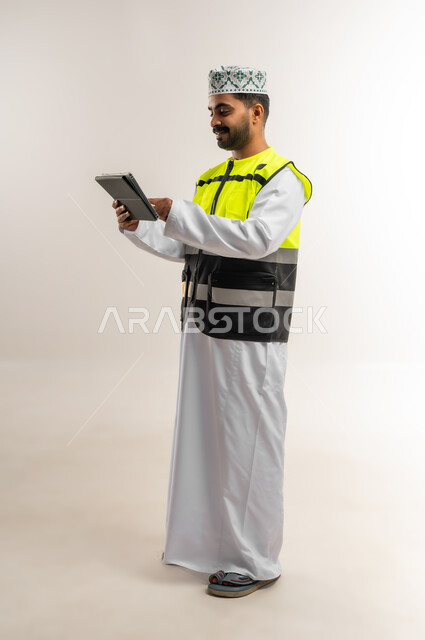 متابعة مخطط الاعمار عبر الجهاز القني, العمل في القطاع الهندسي، مفهوم الهندسة والانشاءات المعمارية، بورتريه لمهندس عربي خليجي عماني يرتدي الدشداشة والكمة وسترة الحماية يمسك بيده التابلت، مهنة ووظيفة للشباب في سلطنة عمان، الجسم بالطول الكامل، خلفية بيضاء