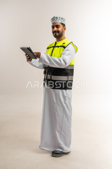 Following up on the reconstruction plan through the technical device, working in the engineering sector, the concept of engineering and architectural construction, a portrait of an Arab Gulf Omani engineer wearing a dishdasha, a keffiyeh, and a protective vest, holding a tablet in his hand, a profession and job for young people in the Sultanate of Oman, full-length body, white background