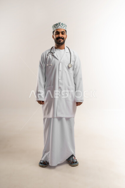 Working in the medical sector in the Sultanate of Oman, the concept of medicine and healthcare, portrait of a smiling Omani Gulf Arab doctor wearing a dishdasha, kummah and professional uniform, holding a stethoscope, standing straight, looking at the camera with self-confident gestures, full body, white background