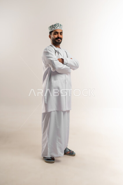 العمل في القطاع الطبي في سلطنة عمان، مفهوم الطب والرعاية الصحية، بورتريه لطبيب عربي خليجي عماني مبتسم يرتدي الدشداشة والكمة والزي الخاص بالمهنة يمسك بيده السماعة الطبية يقف بايدي متقاطعة ينظر إلى الكاميرا يإيماءات الثقة بالنفس، الجسم بالطول الكامل، خلفية بيضاء