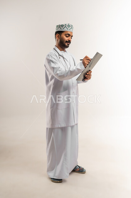العمل في القطاع الطبي في سلطنة عمان، مفهوم الطب والرعاية الصحية، بورتريه لطبيب عربي خليجي عماني مبتسم يرتدي الدشداشة والكمة والزي الخاص بالمهنة يمسك بيده السماعة الطبية بقوم بكتابة وصفة للمريض، الجسم بالطول الكامل، خلفية بيضاء