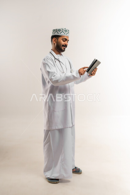 دمج التقنية والتكنولوجيا في الطب والرعاية الصحية، العمل في القطاع الطبي في سلطنة عمان، بورتريه لطبيب عربي خليجي عماني مبتسم يرتدي الدشداشة والكمة والزي الخاص بالمهنة يضع السماعة الطبية يعمل على جهاز التابلت بتعابير السرور والسعادة، الجسم بالطول الكامل، خلفية بيضاء
