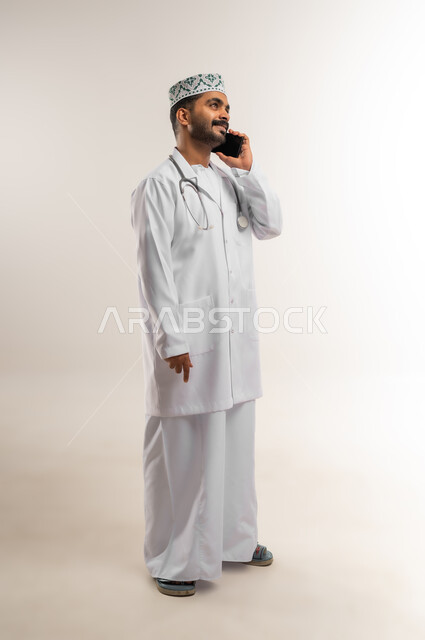 Integrating technology and technology into medicine and healthcare, working in the medical sector in the Sultanate of Oman, portrait of an Arab Gulf Omani doctor wearing a dishdasha, kummah, and professional uniform, wearing a stethoscope, making a phone call via mobile phone with a happy expression, full body, white background