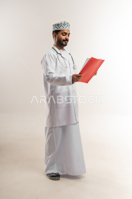 العمل في القطاع الطبي في سلطنة عمان، مفهوم الطب والرعاية الصحية، بورتريه لطبيب عربي خليجي عماني مبتسم يرتدي الدشداشة والكمة والزي الخاص بالمهنة يضع السماعة الطبية يقوم بقراءة سجل المريض، الجسم بالطول الكامل، خلفية بيضاء