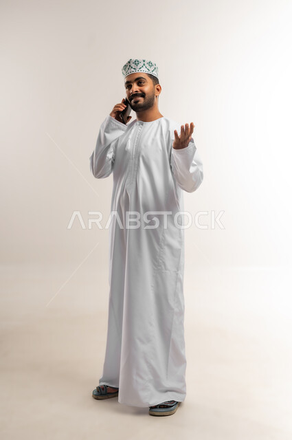 دردشة ومحادثات وتعارف، دمج التكنولوجيا والاجهزة الحديثة في الحياة اليومية، بورتريه لرجل عربي خليجي عماني يرتدي الدشداشه والكمة  يقوم بإجراء مكالمة هاتفية باستخدام الهاتف المحمول، التواصل مع الاهل والاصدقاء، صورة الجسم بالطول الكامل، خلفية بيضاء