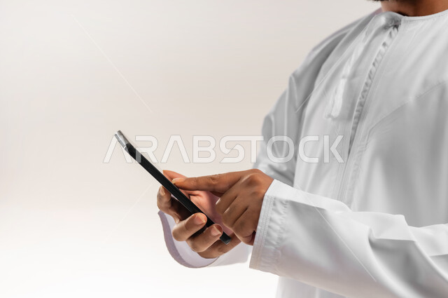 دمج التكنولوجيا في الحياة اليومية، استخدام التقنيات والأجهزة الحديثة، تصفح التطبيقات ومواقع التواصل الاجتماعي عبر الانترنت، دردشة ومحادثات وتعارف، بورتريه مقرب من الخلف لرجل عربي خليجي عماني يرتدي الدشداشة والكمة يقوم باستخدام الهاتف المحمول، خلفية بيضاء