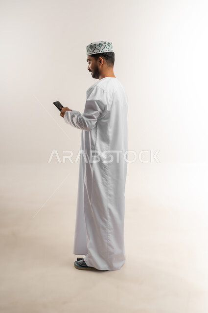 دمج التكنولوجيا في الحياة اليومية، استخدام التقنيات والأجهزة الحديثة، تصفح التطبيقات ومواقع التواصل الاجتماعي عبر الانترنت، دردشة ومحادثات وتعارف، بورتريه لرجل عربي خليجي عماني يرتدي الدشداشة والكمة يقوم باستخدام الهاتف المحمول، الجسم بالطول الكامل، خلفية بيضاء