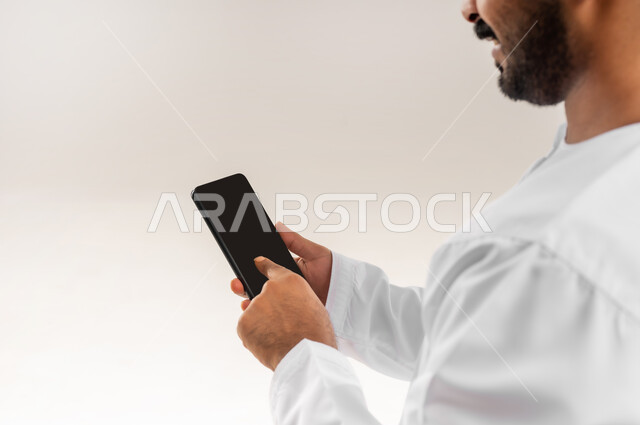 دمج التكنولوجيا في الحياة اليومية، استخدام التقنيات والأجهزة الحديثة، تصفح التطبيقات ومواقع التواصل الاجتماعي عبر الانترنت، دردشة ومحادثات وتعارف، بورتريه مقرب من الخلف لرجل عربي خليجي عماني يرتدي الدشداشة والكمة يقوم باستخدام الهاتف المحمول، خلفية بيضاء