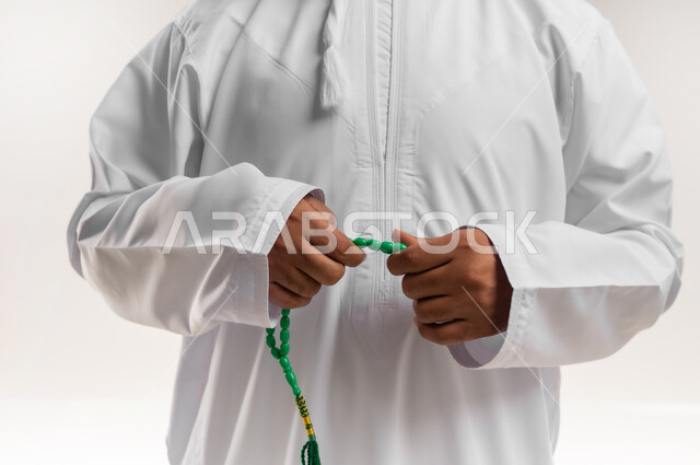 الاستغفار والصلاة على النبي الكريم، مفهوم العبادة والتقرب لرب العالمين، قراءة الأذكار والأدعية، التسبيح والتهليل وذكر الله تعالى، بورتريه مقرب لرجل عربي خليجي عماني يرتدي الدشداشة والكمة يمسك بيده السبحة، خلفية بيضاء