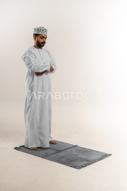 التوجه للقبلة وأداء فريضة الصلاة، التقرب إلى الله بالعبادة والطاعة، بورتريه لرجل عربي خليجي عماني يرتدي الدشداشة والكمه يصلي على السجادة، الحرص على عدم تفويت وقت الصلاة، الجسم بالطول الكامل، خلفية بيضاء