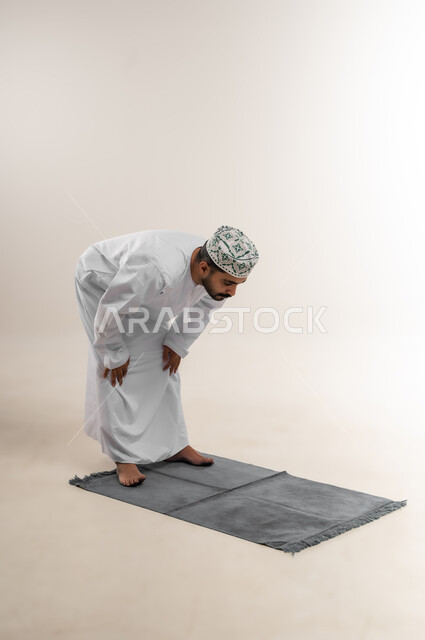 الركوع وأداء فريضة الصلاة، الحرص على عدم تفويت وقت الصلاة، التقرب إلى الله بالعبادة والطاعة، بورتريه لرجل عربي خليجي عماني يرتدي الدشداشة والكمه يقف على السجادة ويركع، خلفية بيضاء