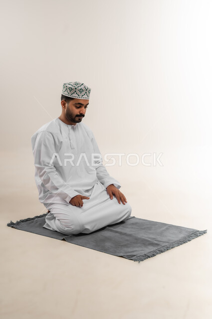 الجلوس للتشهد على السجادة، الخشوع والتضرع إلى الله، بورتريه لرجل عربي خليجي عماني يرتدي الدشداشة والكمه يؤدي فريضة الصلاة، أداء الصلاة في وقتها، التقرب إلى الله بالعبادة والطاعة، خلفية بيضاء