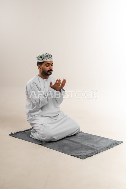 الدعاء بخشوع وتضرع، طلب المغفرة والعفو من الله، بورتريه لرجل عربي خليجي عماني يرتدي الدشداشة والكمه يجلس على سجادة الصلاة يرفع كفيه المفتوحتين للأعلى ويدعو بتذلل، التقرب إلى الله بالعبادة والطاعة، خلفية بيضاء