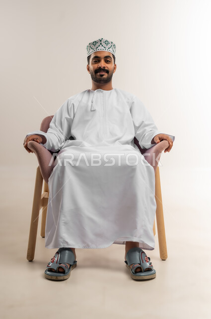 مفهوم الرجولة والوقار، تعابير الثقة بالنفس، الاهتمام بالمظهر الخارجي، بورتريه لرجل عربي خليجي عماني يرتدي الدشداشة والكمة يجلس على كرسي مريح ينظر الى الكاميرا بايماءات الجدية، خلفية بيضاء