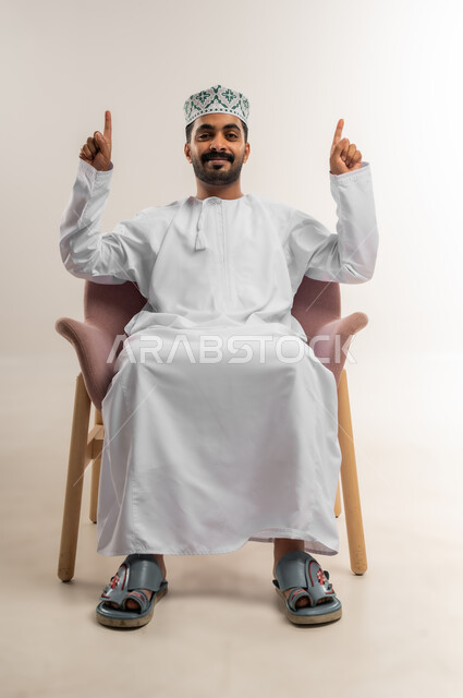 تحديد موقع شيء ما، اشارات يدين تدل على هذا المكان (هنا)، بورتريه لرجل عربي خليجي عماني مبتسم يرتدي الدشداشة والكمة يجلس على كرسي مريح يشير بأصبعي السبابة بايماءات السرور والسعادة، خلفية بيضاء