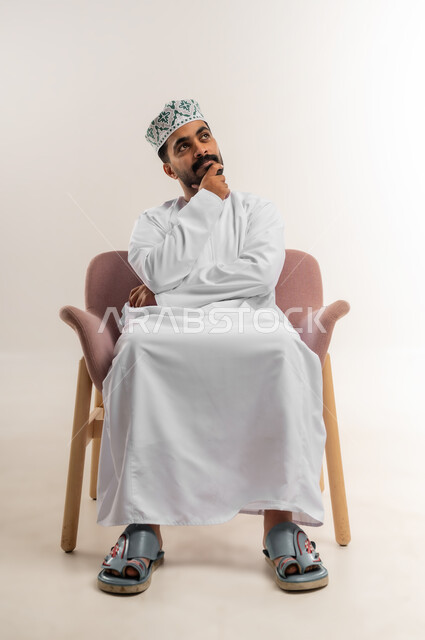 ايجاد حل لمشكلة ما، التأمل والتخطيط للمستقبل، بورتريه لرجل عربي خليجي عماني يرتدي الدشداشة والكمة يضع يده على ذقنه ينظر الى مكان ما بايماءات التفكير والتركيز، تعابير شرود الذهن والتمعن، خلفية بيضاء