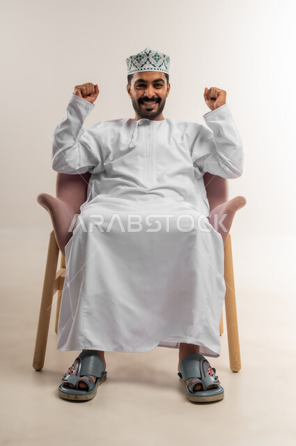القوة والتحدي والحماس، مفهوم الانجاز وتحقيق الاهداف، بورتريه لرجل عربي خليجي عماني يرتدي الدشداشة والكمة يجلس على كرسي مريح يرفع قبضتيه نحو الأعلى بإيماءات التشجيع، تعابير السرور والسعادة، خلفية بيضاء
