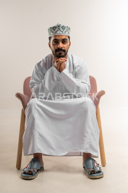 Concern for elegance and outward appearance, the concept of strength, masculinity and chic, a portrait of a smiling Omani Gulf Arab man wearing a dishdasha and a kumrah sitting on a comfortable chair with clasped hands looking at the camera with expressions of joy and pleasure, gestures of self-confidence, white background