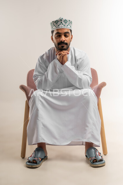 Concern for elegance and outward appearance, the concept of strength, masculinity and chic, a portrait of a smiling Omani Gulf Arab man wearing a dishdasha and a kumrah sitting on a comfortable chair with clasped hands looking at the camera with expressions of joy and pleasure, gestures of self-confidence, white background