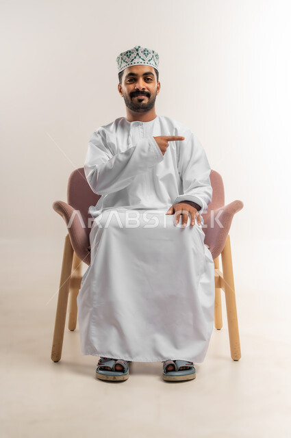 تحديد موقع شيء ما، اشارات يدين تدل على هذا المكان (هنا)، بورتريه لرجل عربي خليجي عماني مبتسم يرتدي الدشداشة والكمة يجلس على كرسي مريح يشير بأصبعي السبابة بايماءات السرور والسعادة، خلفية بيضاء