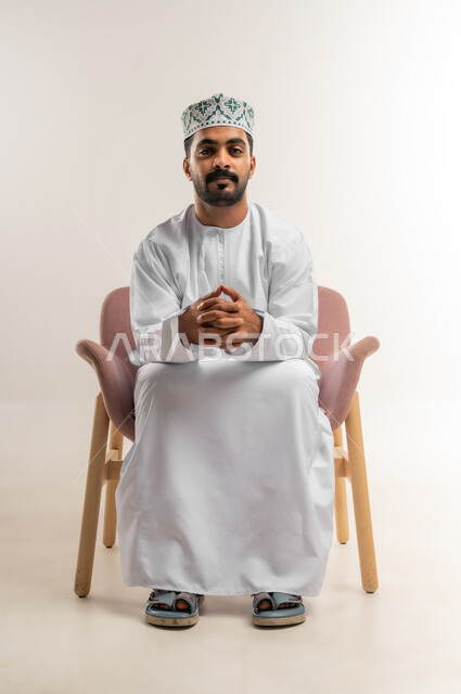 Concern for elegance and outward appearance, the concept of strength, masculinity and chic, a portrait of a smiling Omani Gulf Arab man wearing a dishdasha and a kumrah sitting on a comfortable chair with clasped hands looking at the camera with expressions of joy and pleasure, gestures of self-confidence, white background