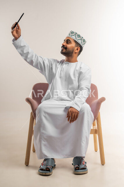 Integrating technology and modern devices into daily life, portrait of a smiling Omani Gulf Arab man wearing a dishdasha and a kuma sitting on a chair taking souvenir selfies using a mobile phone, communicating with family and friends, white background
