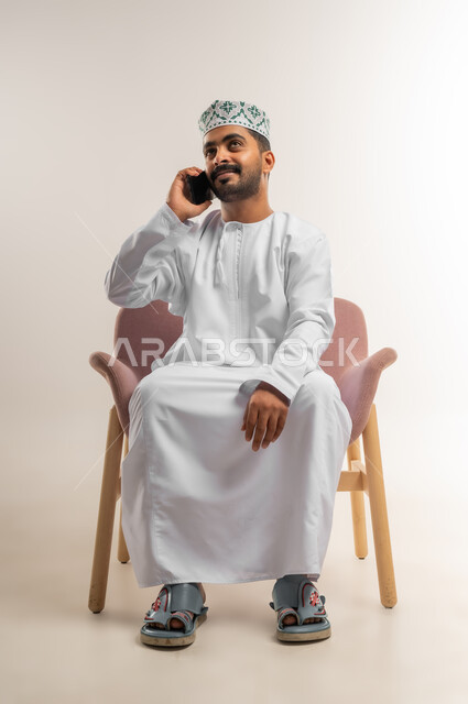 دمج التكنولوجيا والاجهزة الحديثة في الحياة اليومية، بورتريه لرجل عربي خليجي عماني يرتدي الدشداشه والكمة يجلس على كرسي مريح يقوم بإجراء مكالمة هاتفية باستخدام الهاتف المحمول، التواصل مع الاهل والاصدقاء، دردشة ومحادثات وتعارف، خلفية بيضاء