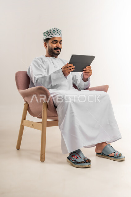 الدردشة مع الأهل والأصدقاء، استعمال التكنولوجيا والتقنيات الحديثة، بورتريه لرجل عربي خليجي عماني يرتدي الدشداشة والكمة يحمل بيده جهاز التابلت، تصفح مواقع التواصل الاجتماعي، استخدام تطبيقات وبرامج الأجهزة اللوحية، خلفية بيضاء