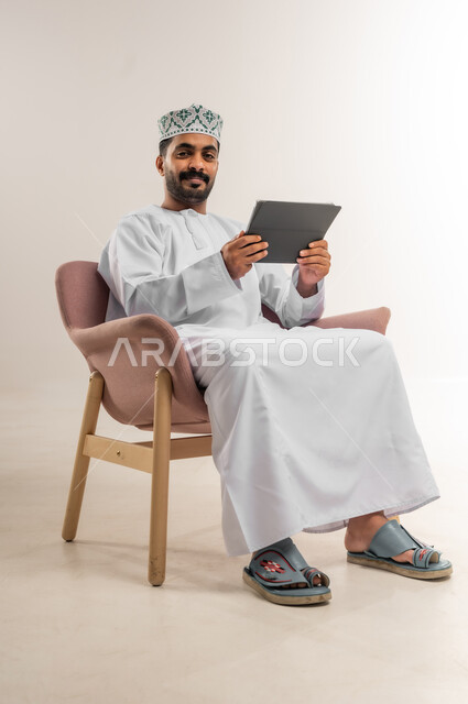 الدردشة مع الأهل والأصدقاء، استعمال التكنولوجيا والتقنيات الحديثة، بورتريه لرجل عربي خليجي عماني يرتدي الدشداشة والكمة يحمل بيده جهاز التابلت، تصفح مواقع التواصل الاجتماعي، استخدام تطبيقات وبرامج الأجهزة اللوحية، خلفية بيضاء