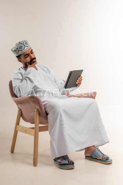 الدردشة مع الأهل والأصدقاء، استعمال التكنولوجيا والتقنيات الحديثة، بورتريه لرجل عربي خليجي عماني يرتدي الدشداشة والكمة يحمل بيده جهاز التابلت، تصفح مواقع التواصل الاجتماعي، استخدام تطبيقات وبرامج الأجهزة اللوحية، خلفية بيضاء