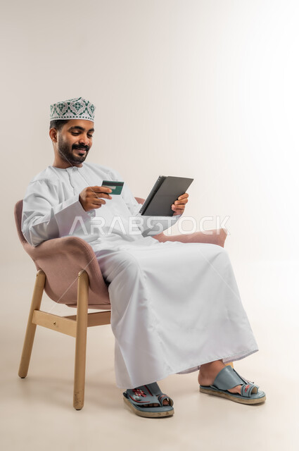 الدفع الإلكتروني عبر التقنيات الحديثة المتطورة، استخدام تطبيقات الجهاز اللوحي للدفع والشراء، بورتريه لشاب عربي خليجي عماني يرتدي الدشداشة والكمة يجلس على كرسي يمسك بيده البطاقة الإلكترونية والتابلت, خلفية بيضاء