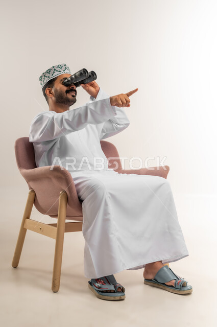 حب الاستكشاف والمغامرة، رؤية الاشياء البعيدة وتقريبها، بورتريه لرجل عربي خليجي عماني يرتدي الدشداشة والكمة يجلس على كرسي مريح يحمل بيده منظار ثنائي العينين وينظر بإيماءات السرور والاستمتاع، خلفية بيضاء
