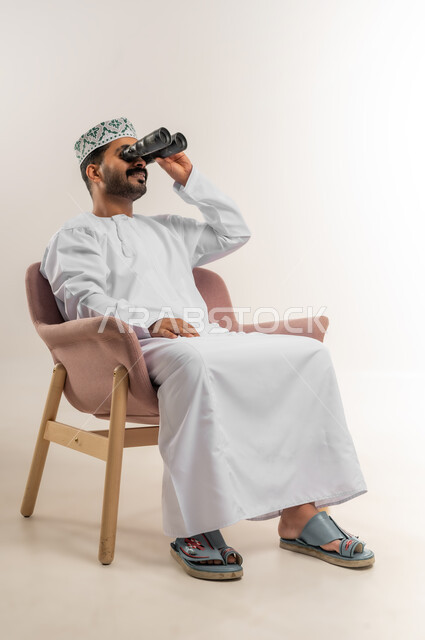 حب الاستكشاف والمغامرة، رؤية الاشياء البعيدة وتقريبها، بورتريه لرجل عربي خليجي عماني يرتدي الدشداشة والكمة يجلس على كرسي مريح يحمل بيده منظار ثنائي العينين وينظر بإيماءات السرور والاستمتاع، خلفية بيضاء