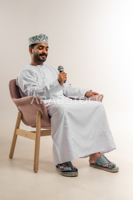 العمل في تقديم الفقرات و البرامج الفنية الترفيهية, تعابير وإيماءات السعادة, بورتريه لشاب عربي خليجي عماني يرتدي الدشداشة و الكمة يجلس على كرسي مريح يمسك بيده المايكروفون، الاستمتاع بالغناء وتقديم المواهب الفنية، خلفية بيضاء