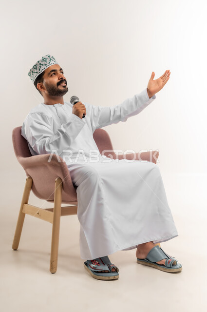 العمل في تقديم الفقرات و البرامج الفنية الترفيهية, تعابير وإيماءات السعادة, بورتريه لشاب عربي خليجي عماني يرتدي الدشداشة و الكمة يجلس على كرسي مريح يمسك بيده المايكروفون، الاستمتاع بالغناء وتقديم المواهب الفنية، خلفية بيضاء